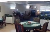 A common area in a residential care home featuring several round tables covered with green tablecloths and floral centerpieces. The room has multiple chairs around the tables, potted plants, and a seating area with armchairs and a television in the background. The walls are light-colored with windows letting in natural light.