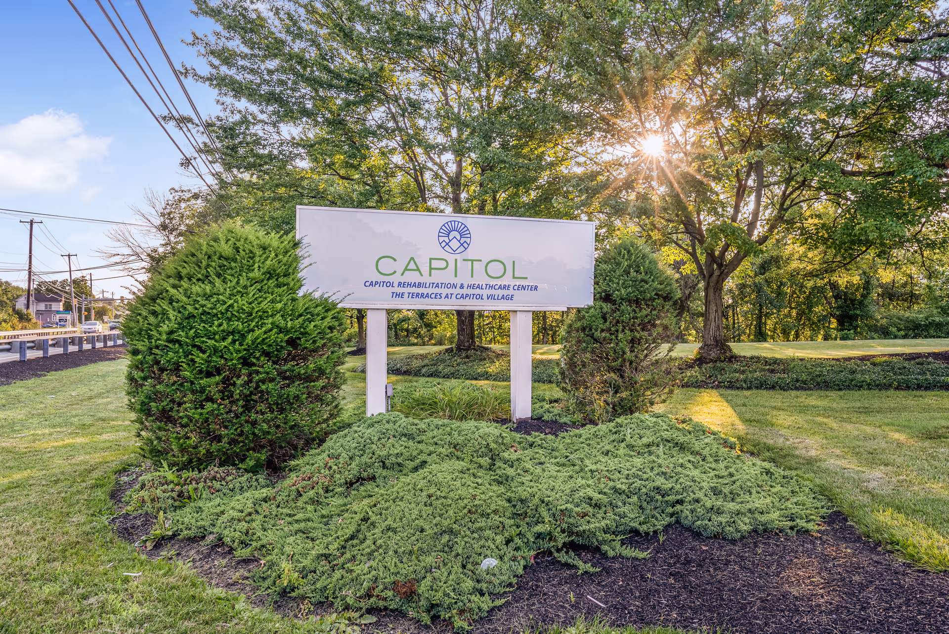 Outdoor view of a landscaped area with green bushes and trees surrounding a white sign that reads 'CAPITOL Rehabilitation & Healthcare Center The Terraces at Capitol Village' with the sun shining through the trees in the background.