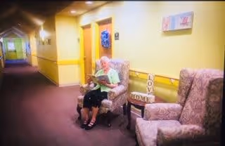 An elderly woman sitting in a cushioned armchair in a hallway of an assisted living facility. The hallway has yellow walls with a chair rail, carpeted floor, and several doors. Next to the woman is a small table with decorative blocks spelling 'LOVE' and 'HOME'. Another cushioned armchair is nearby, and a colorful artwork hangs on the wall.
