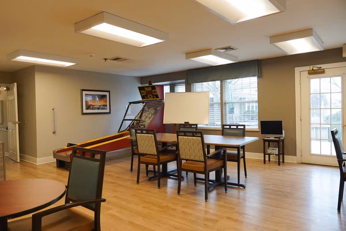 A well-lit room with wooden flooring featuring a table surrounded by chairs, a basketball arcade game, a whiteboard, and large windows with blinds. There is also a door with glass panes leading outside and a small table with a computer monitor on it.