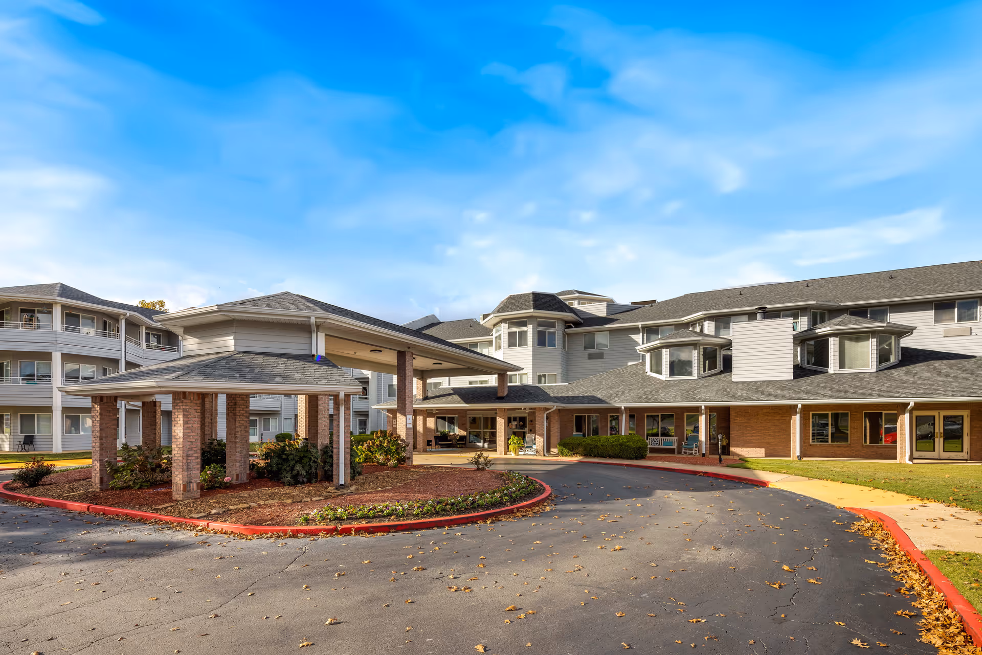 Exterior view of Jackson Meadow - A Provincial Senior Living Community, showing a large multi-story building with a covered entrance driveway, landscaped areas with shrubs, and a clear blue sky.