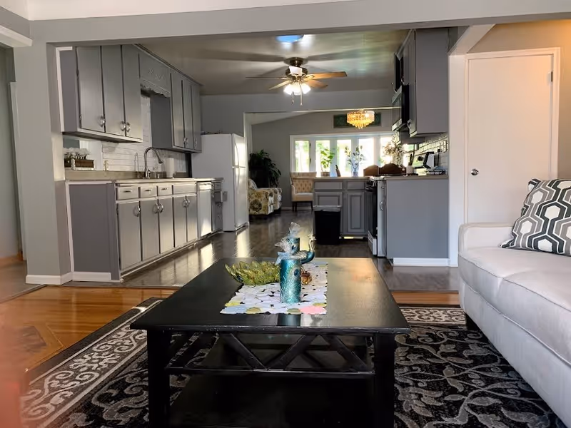 View of a modern open-concept living space featuring a gray kitchen with cabinets, a white refrigerator, and stainless steel appliances. In the foreground, there is a black coffee table with decorative items on a patterned rug, and a light-colored sofa with a geometric patterned pillow. The background shows a dining area with chairs and large windows letting in natural light.