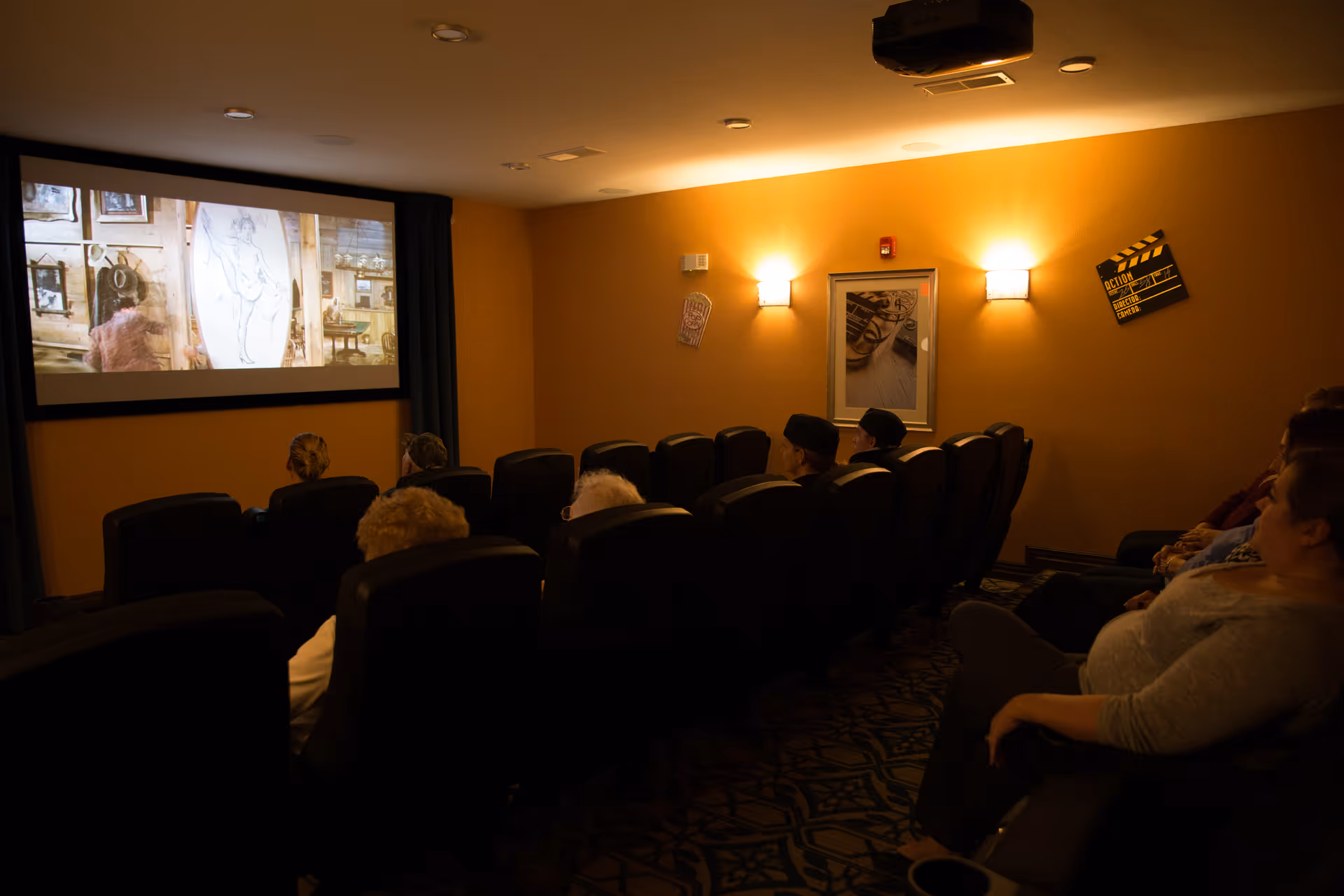 A small movie theater room with several rows of black reclining chairs occupied by elderly people watching a film projected on a large screen. The walls are painted warm yellow with framed pictures and movie-themed decorations. The room is dimly lit by wall sconces.
