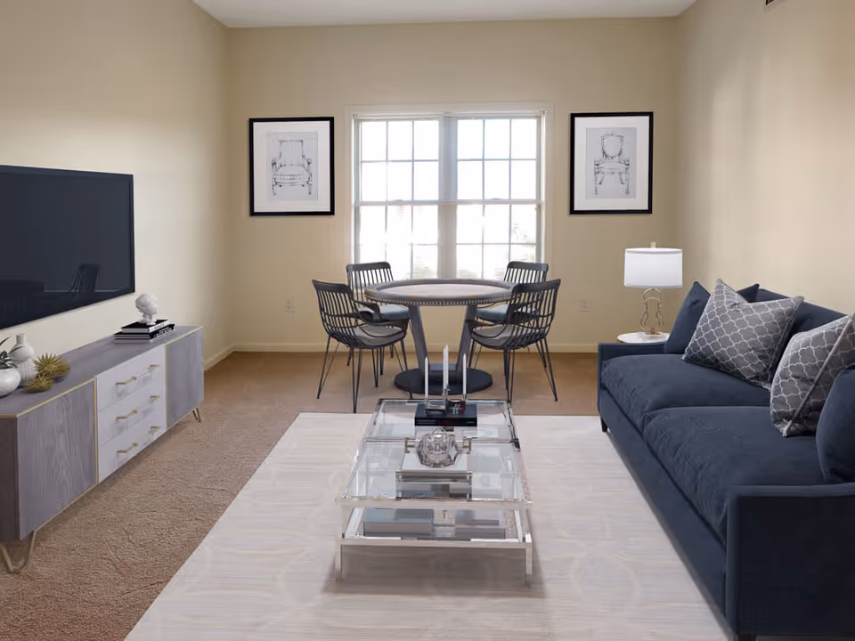 A cozy living room featuring a dark blue sofa, a glass coffee table, and a dining area with a round table and chairs. The walls are painted a soft beige, and there are framed artworks on the walls.