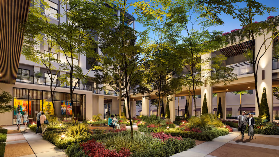 Outdoor courtyard area of Belmont Village Senior Living Coral Gables with landscaped gardens, trees, and walking paths. Several people are seen walking and sitting on benches, surrounded by modern building structures with balconies and large windows.