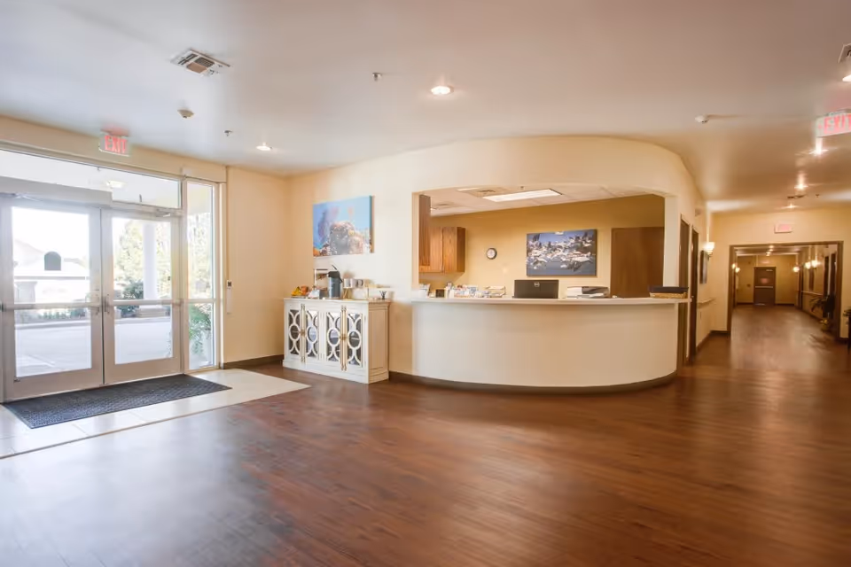 Bright and spacious reception area with wooden flooring, a curved white reception desk, and a glass entrance door letting in natural light. There is a white cabinet with decorative circular mirrors on the left side of the desk, and artwork on the walls. A hallway extends to the right of the reception desk.