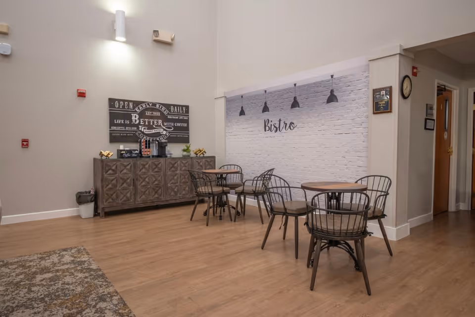 A cozy bistro area inside a senior living facility with wooden floors, a white brick accent wall with the word 'Bistro' written on it, three round wooden tables each surrounded by four metal chairs with cushions, and a wooden cabinet against the wall holding a coffee machine and coffee supplies. The space is well-lit with wall-mounted lights and has a clock and framed pictures on the adjacent wall.
