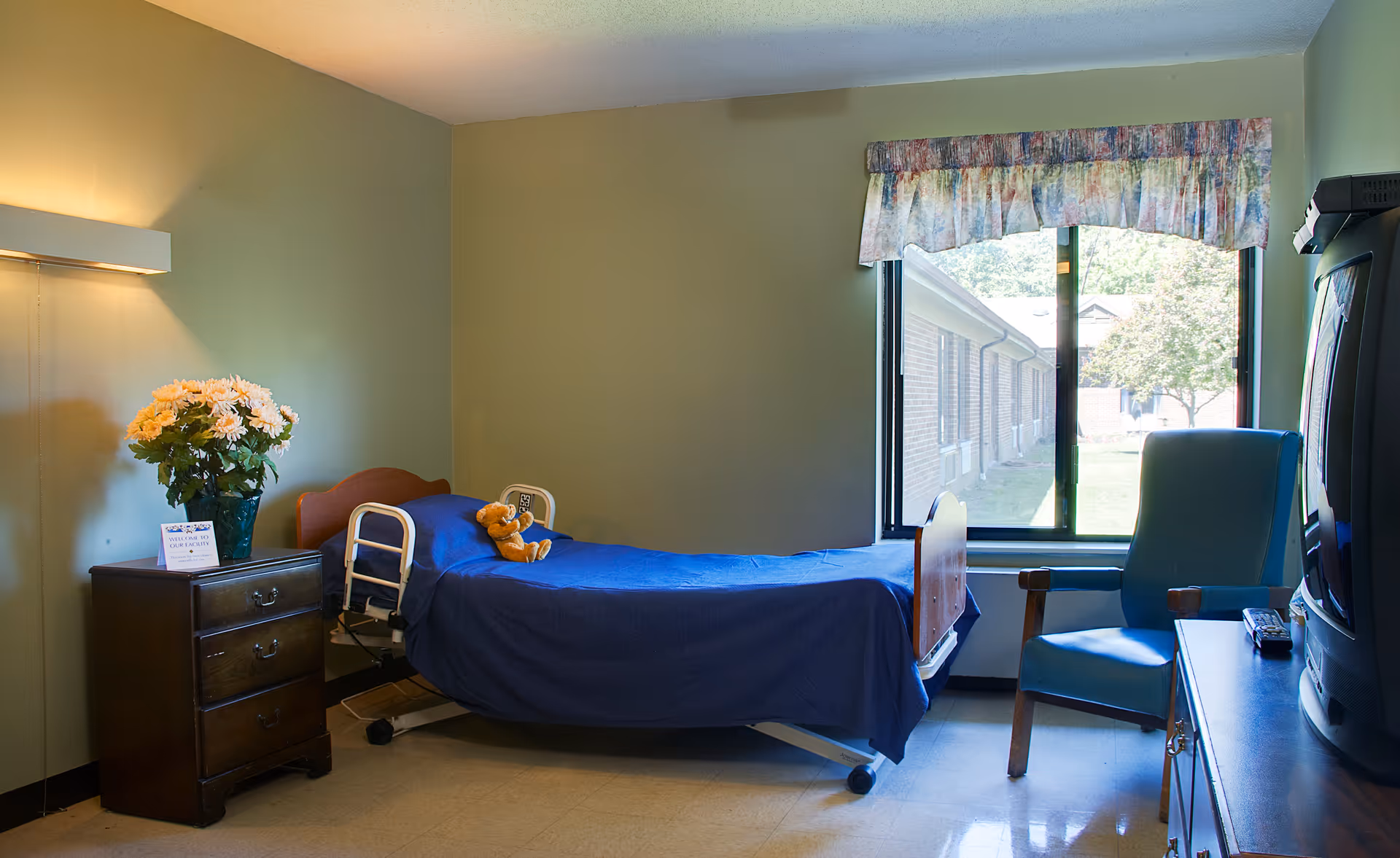 A simple bedroom in a senior living facility with a single bed covered in a blue blanket and a teddy bear on it. Next to the bed is a wooden nightstand with a vase of flowers and a small sign. A large window with a floral valance lets in natural light. There is a blue armchair and a television on a stand opposite the bed.
