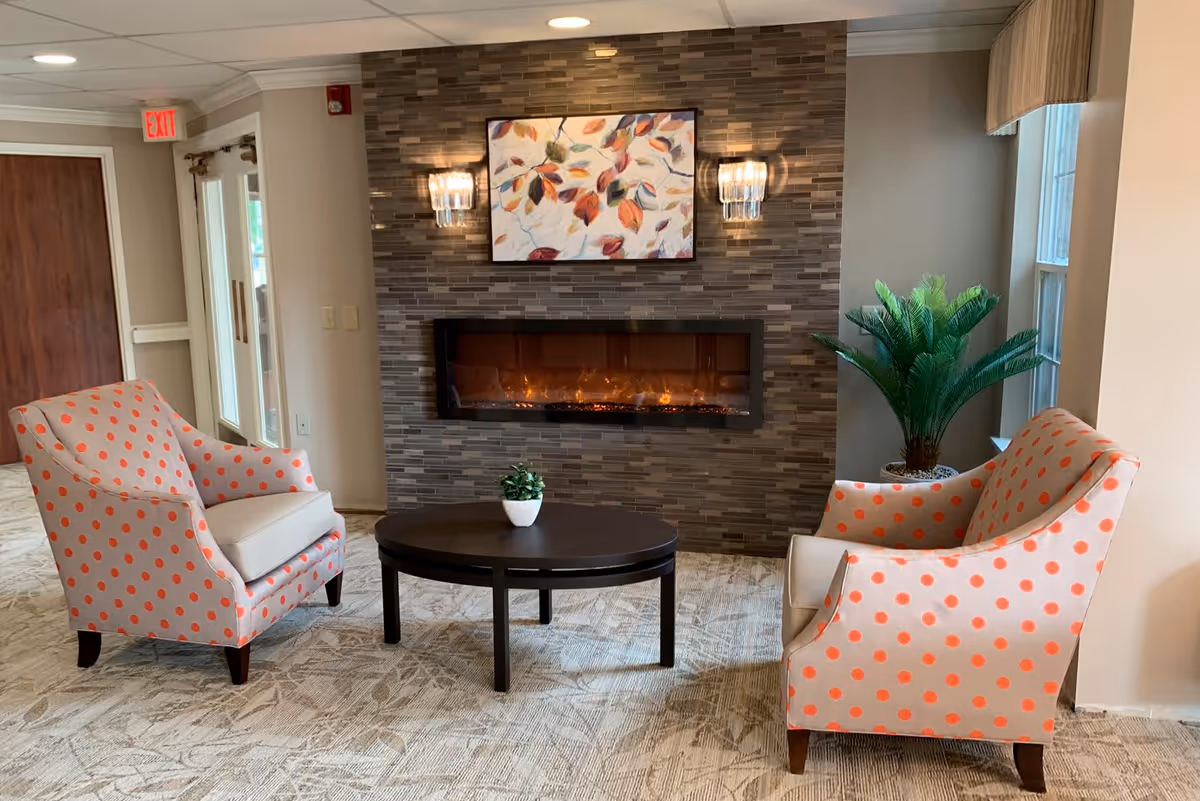 A cozy seating area in a retirement facility with two beige armchairs featuring orange polka dots facing a dark round coffee table with a small potted plant. Behind the table is a modern electric fireplace set in a tiled wall with a colorful leaf-themed painting above it and two wall sconces on either side. A large green plant is placed near a window on the right side, and an exit sign is visible near a door on the left.