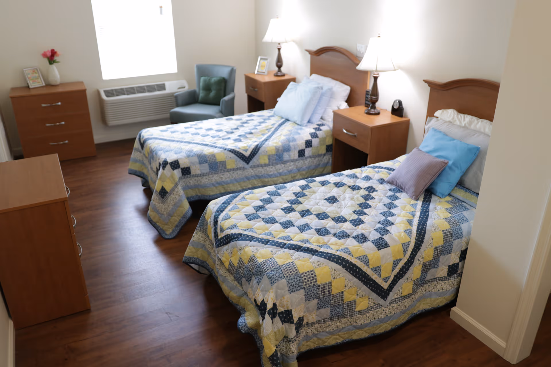 Bright, simply furnished bedroom with two twin beds, nightstands, lamps, a dresser and a chair.