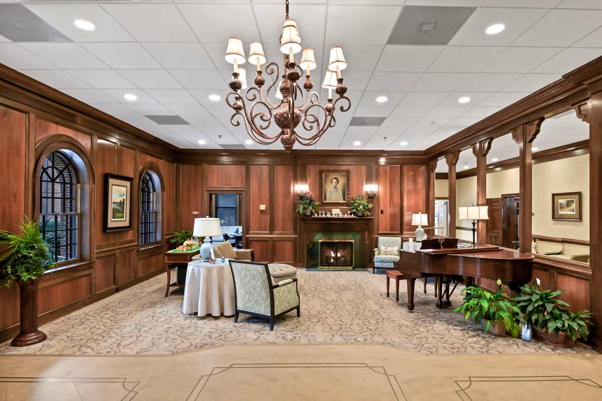 A spacious, elegant living room with wood-paneled walls and a large chandelier hanging from the ceiling. The room features a fireplace with a portrait above it, a grand piano on the right side, several armchairs, a round table with a lamp, and various plants. Large arched windows are on the left wall, and the carpet has a subtle floral pattern.