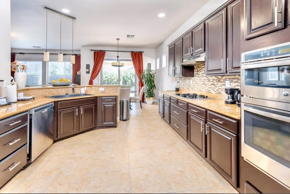 Bright modern kitchen with dark wood cabinets, granite countertops, stainless steel appliances, tile floor, and a breakfast bar opening to a dining area with red curtains.