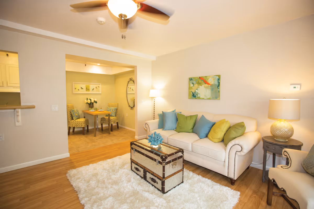 A cozy living room with a white sofa adorned with blue and green pillows, a vintage trunk coffee table on a fluffy white rug, a side table with a lamp, and a ceiling fan. In the background, there is a small dining area with a table, two patterned chairs, a vase with flowers, a round mirror, and framed artwork on the wall.