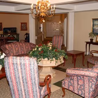 A furnished common living area with patterned upholstered chairs around a central planter-topped table, a chandelier overhead, and a hallway leading to double doors.