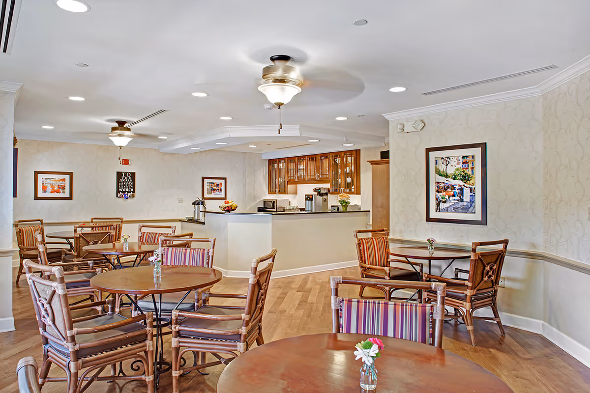 Bright communal dining area with round tables and striped chairs, a serving counter and framed artwork on the walls.