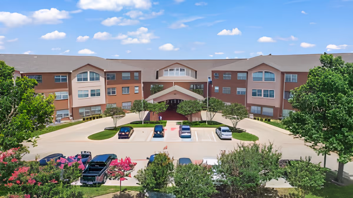 Front exterior of a three-story senior living facility with a porte-cochère, parking lot and landscaped trees.