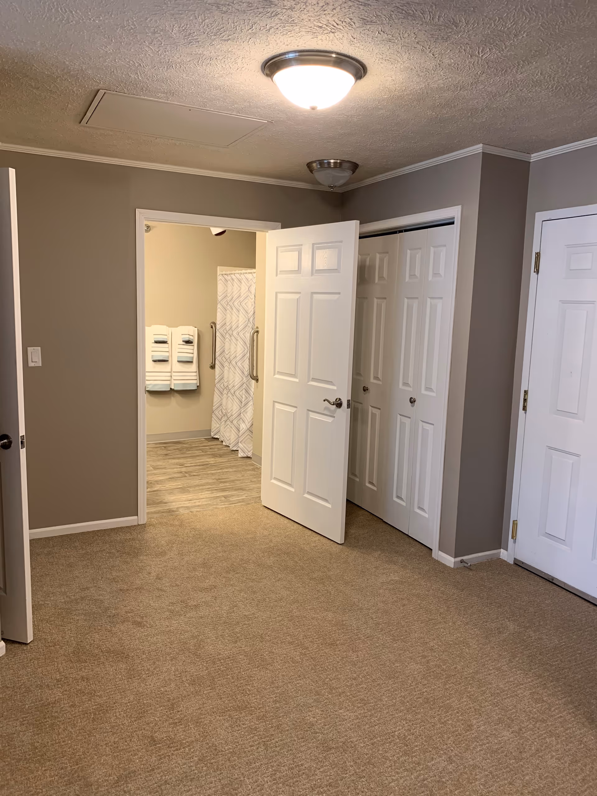 Carpeted bedroom interior with an open door leading to an adjoining bathroom showing towels, grab bars, and a shower curtain.