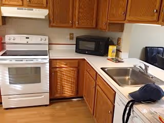Interior view of a kitchen in an assisted living facility featuring a white stove with oven, wooden cabinets, a microwave on the countertop, a stainless steel sink, and various kitchen items including a hat and a bag on the counter.