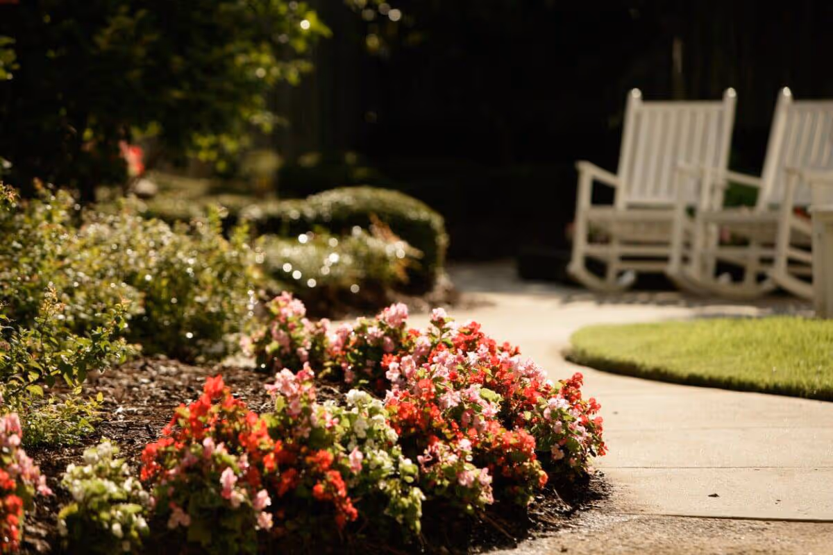 A garden area with colorful flowers in the foreground and a curved concrete pathway leading to white wooden rocking chairs in the background, surrounded by greenery.