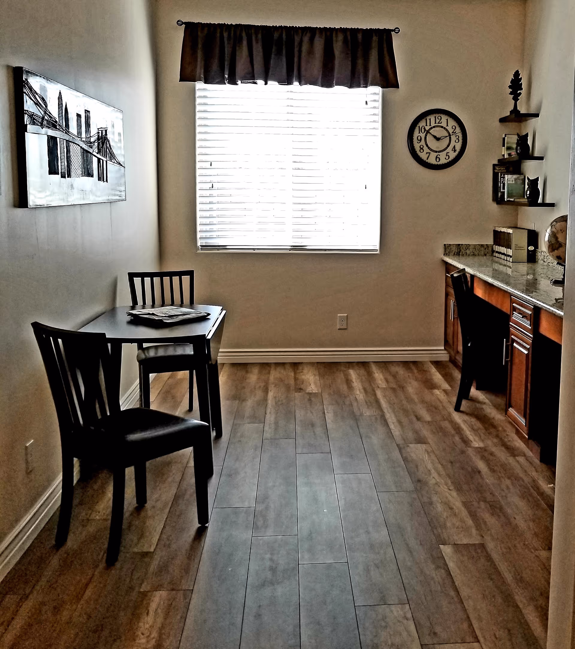 A small room with wooden flooring featuring a table with two chairs on the left side and a built-in desk with a chair on the right side. The desk has shelves with books and decorative items, and a globe is placed on the countertop. A window with blinds and a dark valance is centered on the back wall, and a round wall clock is mounted to the right of the window. A black and white artwork of a bridge is hung on the left wall.