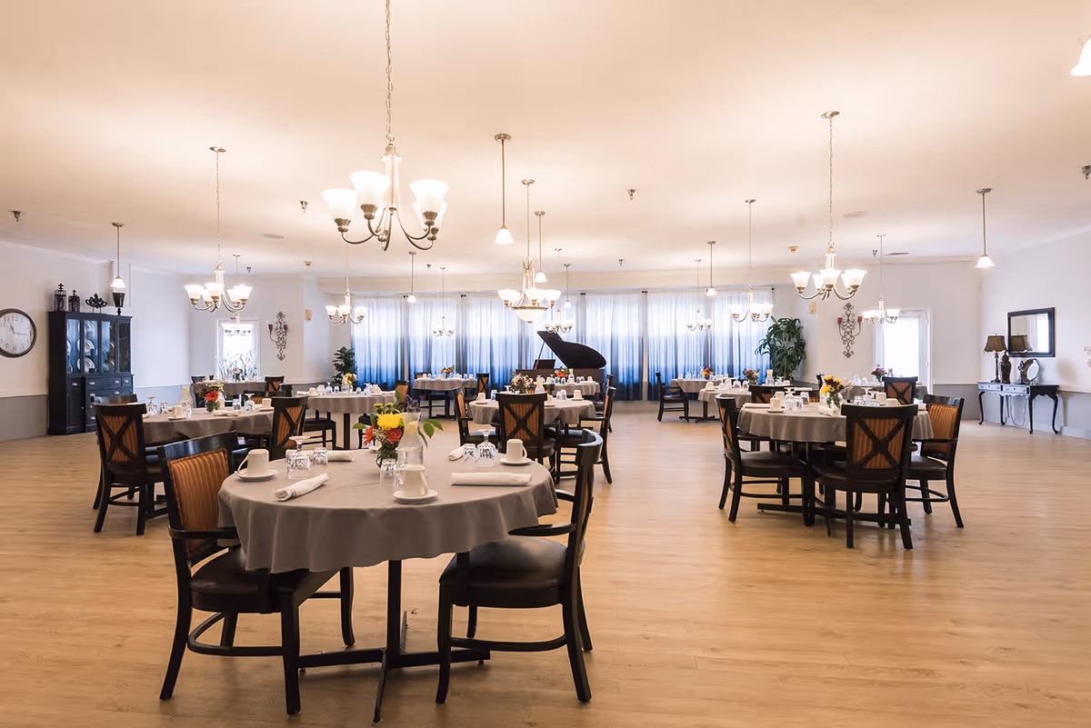 Spacious dining room with round tables set for meals, chandeliers overhead, and a grand piano in the background.