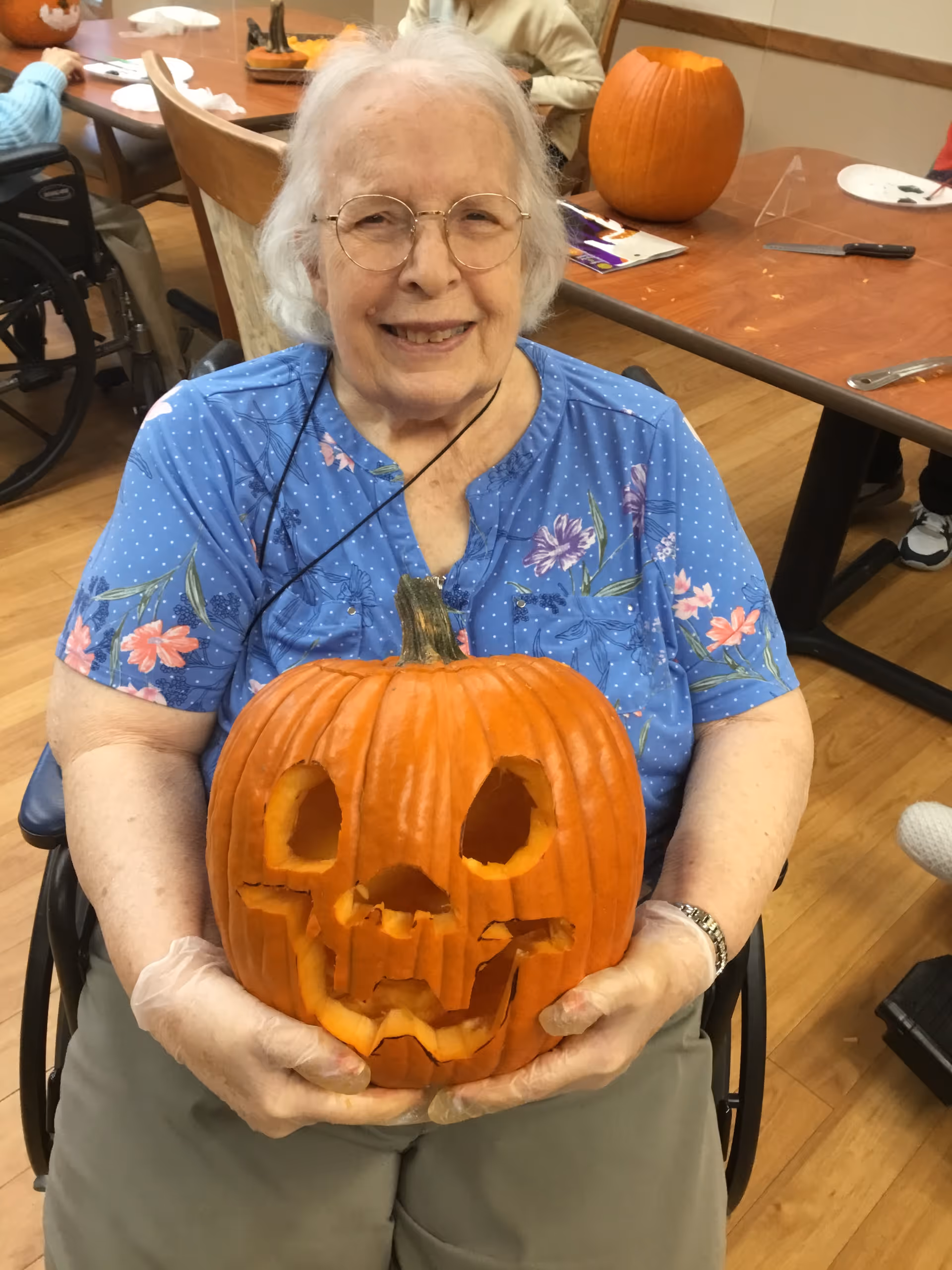 An elderly woman sitting in a wheelchair indoors, smiling and holding a carved Halloween pumpkin with a face. She is wearing glasses, a blue floral shirt, and disposable gloves. In the background, there are other people, tables, and another pumpkin on the table.