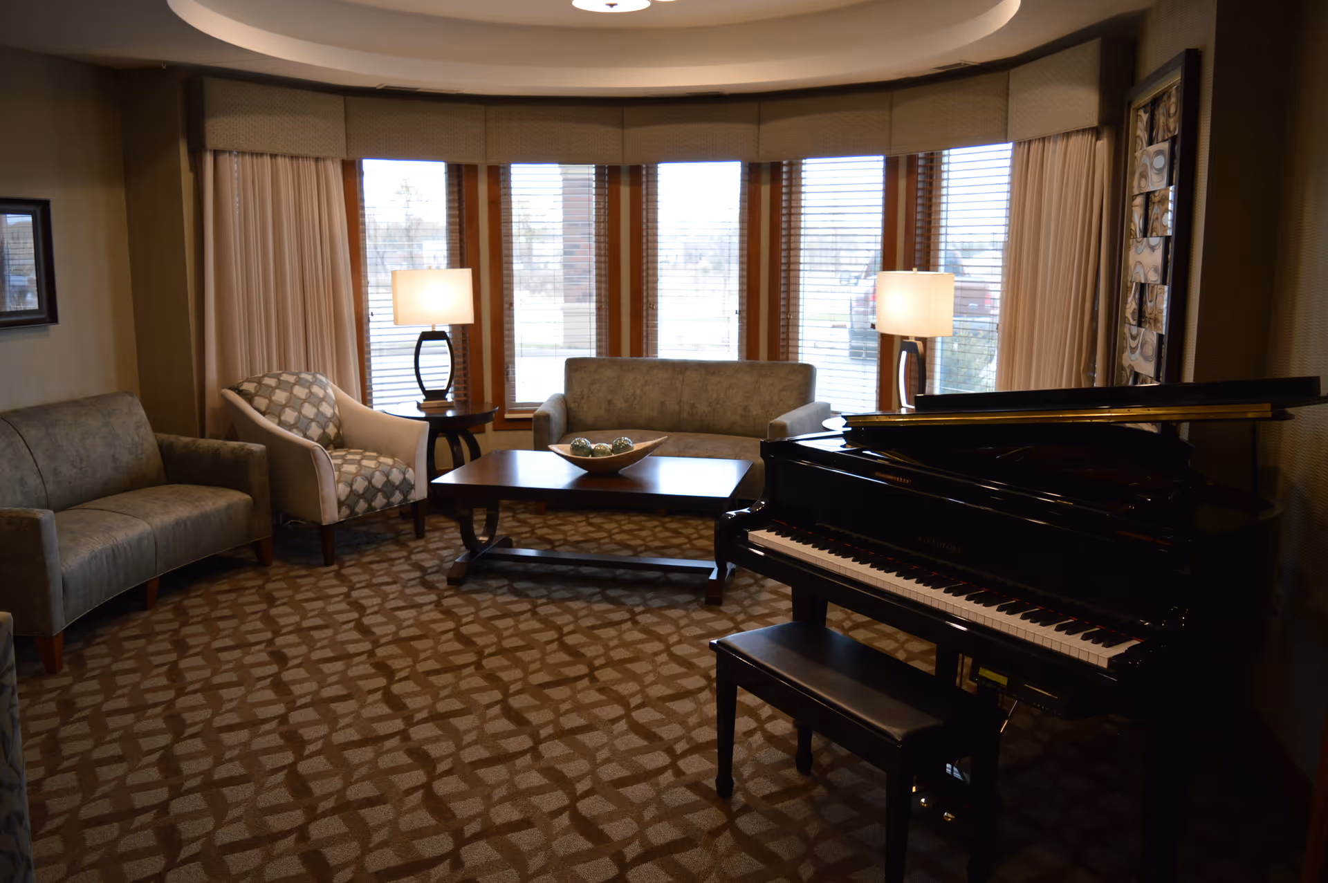 A cozy living room with a grand piano on the right, two sofas and an armchair arranged around a wooden coffee table, two table lamps, and large windows with blinds and curtains letting in natural light.