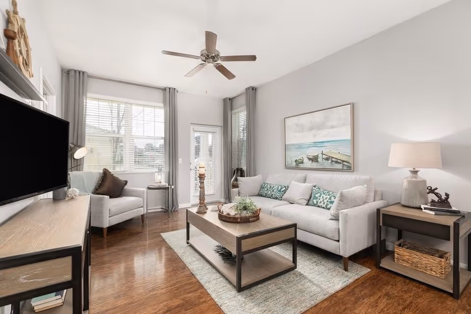 Bright living room with a light-gray sofa, coffee table, armchair, wall-mounted TV, and coastal artwork.