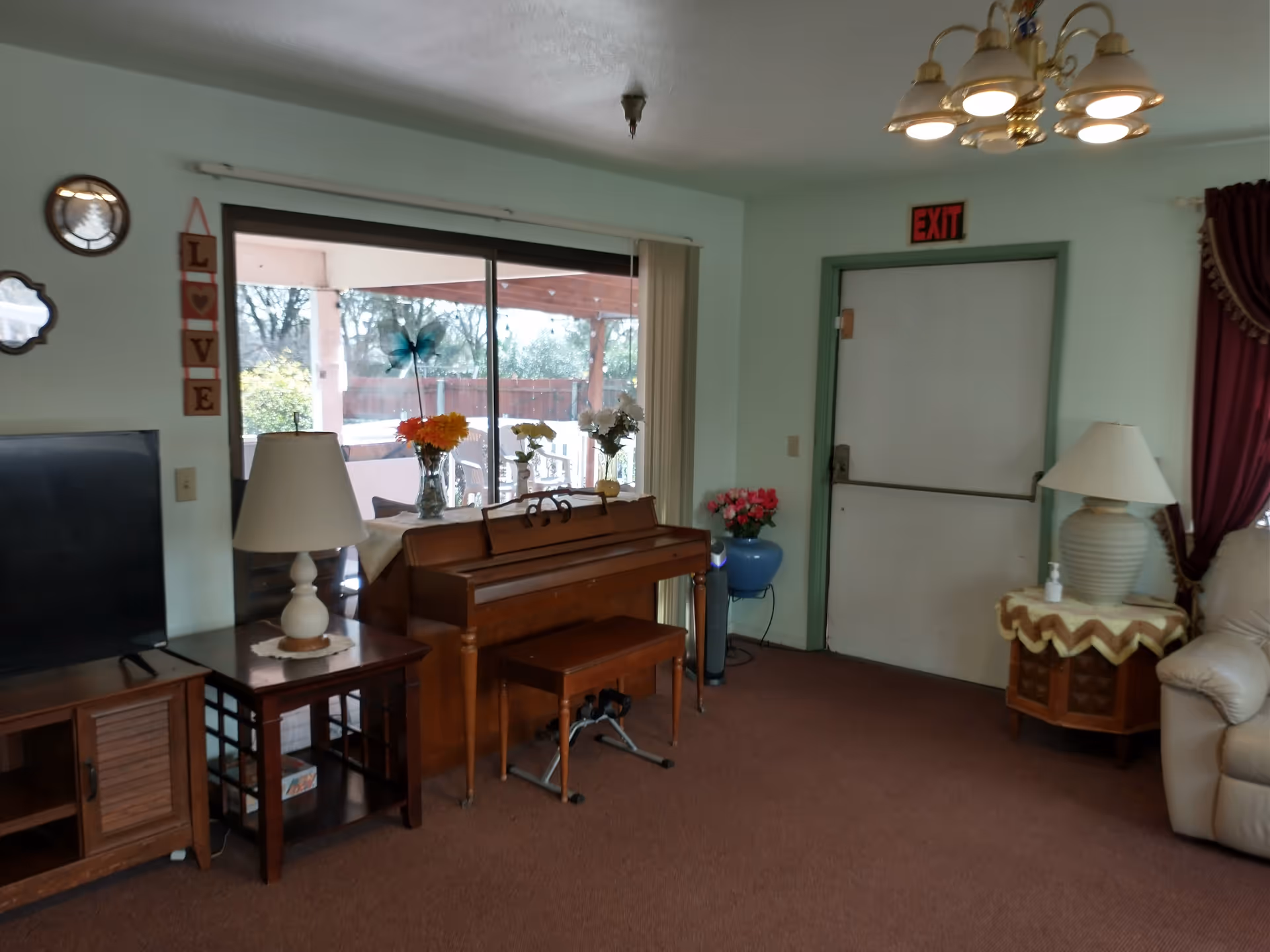 A cozy living room area with a wooden piano and bench, a TV on a wooden stand, two table lamps on side tables, a sliding glass door leading to an outdoor patio, and a white exit door with a red exit sign above it. The room has light green walls, brown carpet, and decorative flowers on the piano and in a blue vase near the door.