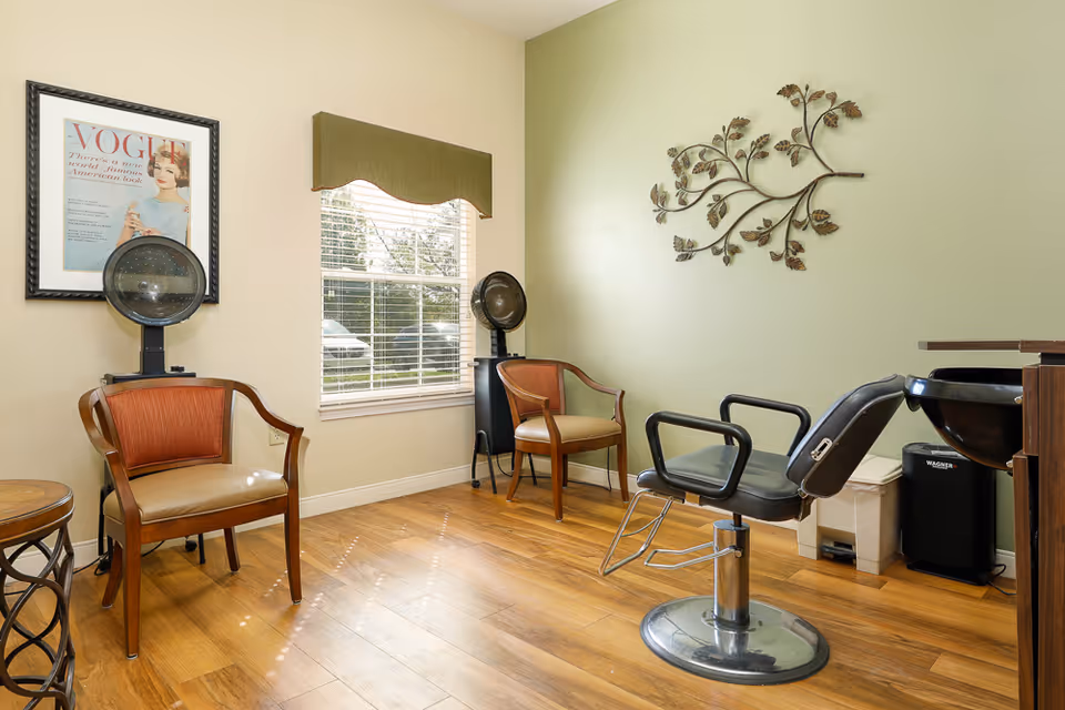 A small salon area with two vintage hair dryers, two wooden chairs with tan cushions, a black salon chair in front of a black wash basin, a window with blinds and a green valance, a framed Vogue magazine cover on the wall, and a decorative metal wall art shaped like a branch with leaves.