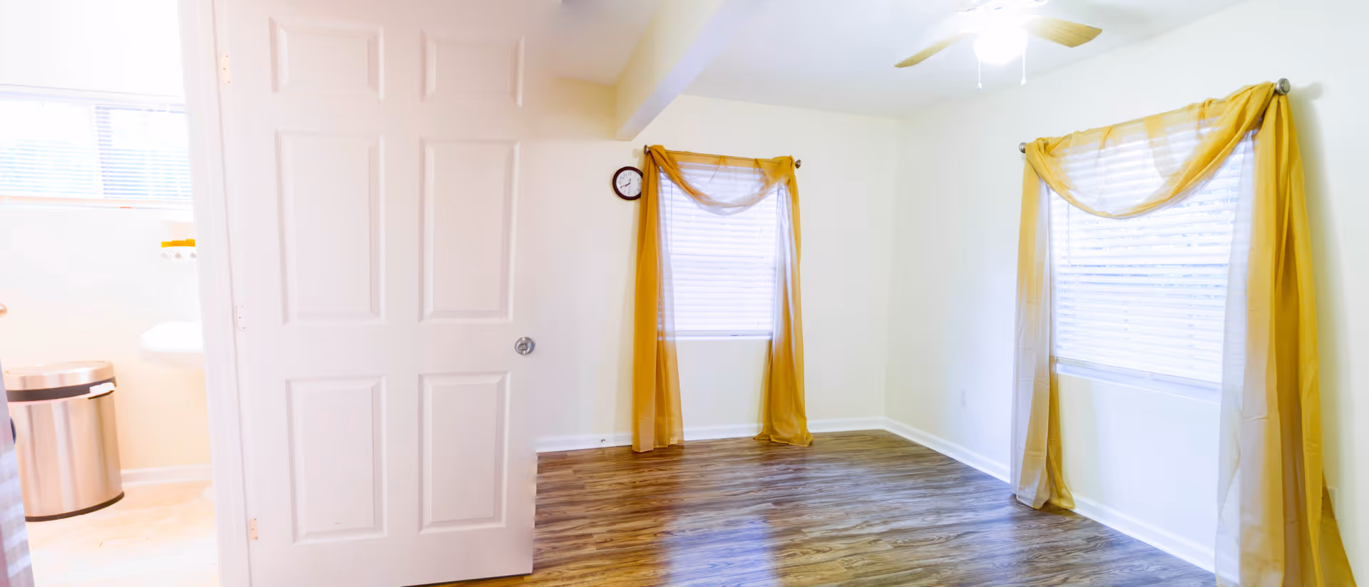 A bright room with wooden flooring and two windows covered with sheer yellow curtains. A white door is partially open, revealing a bathroom with a trash can and a sink. A ceiling fan with a light is visible on the ceiling.