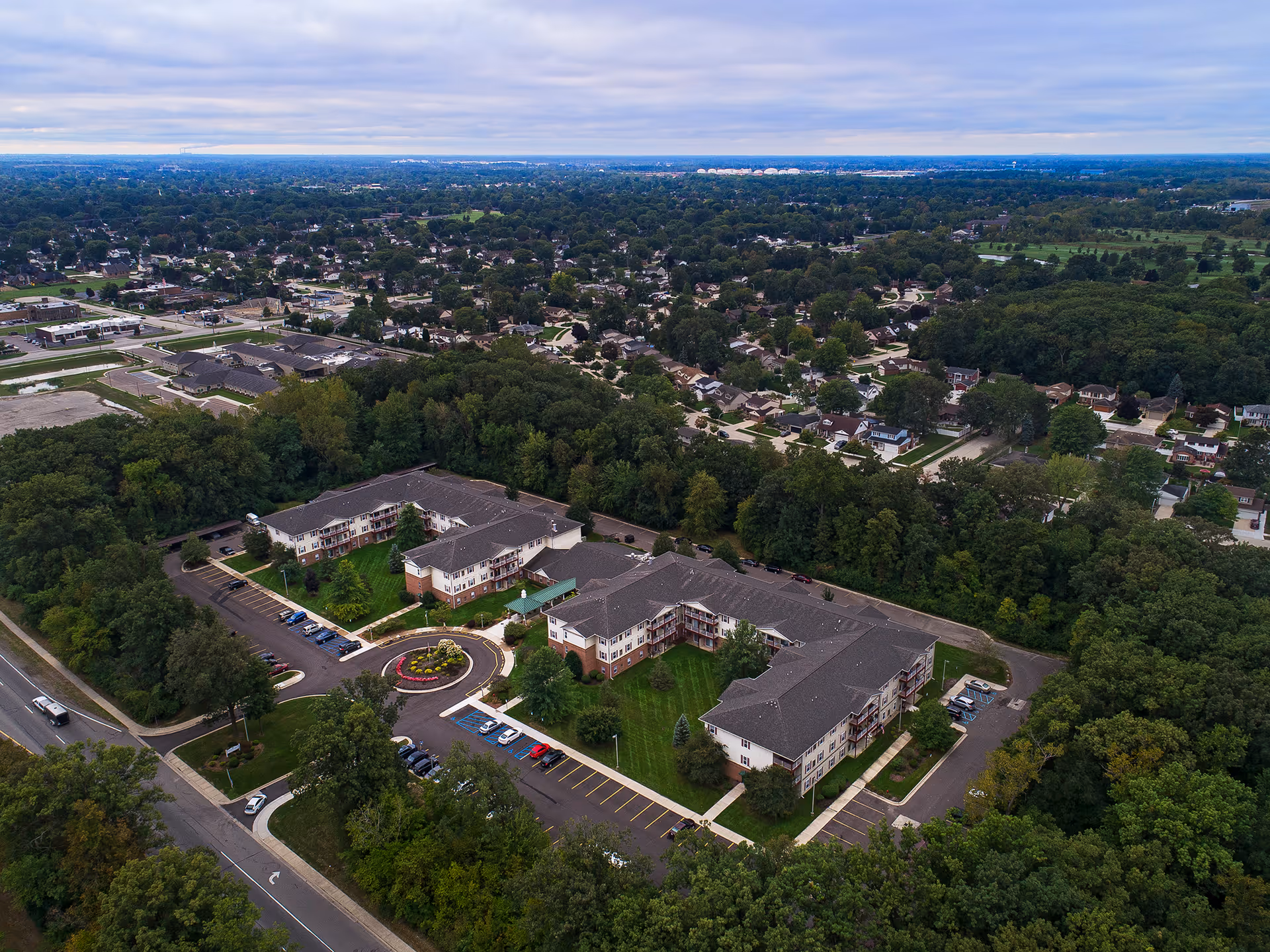Aerial view of a multi-building senior living complex surrounded by trees, parking lots, and a nearby residential neighborhood.