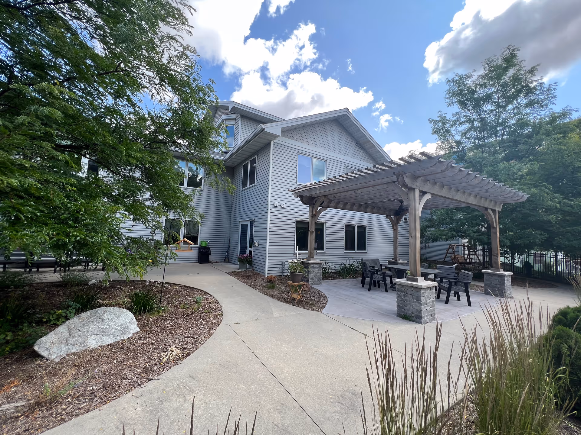 Two-story gray-siding building with a paved courtyard and a wooden pergola with seating under a partly cloudy sky.