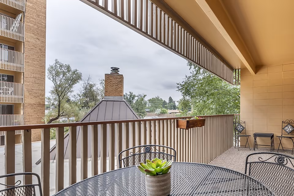Covered balcony area with a metal table and chairs, a small potted plant on the table, railing with a flower box, and a view of trees and a neighboring building under a cloudy sky.