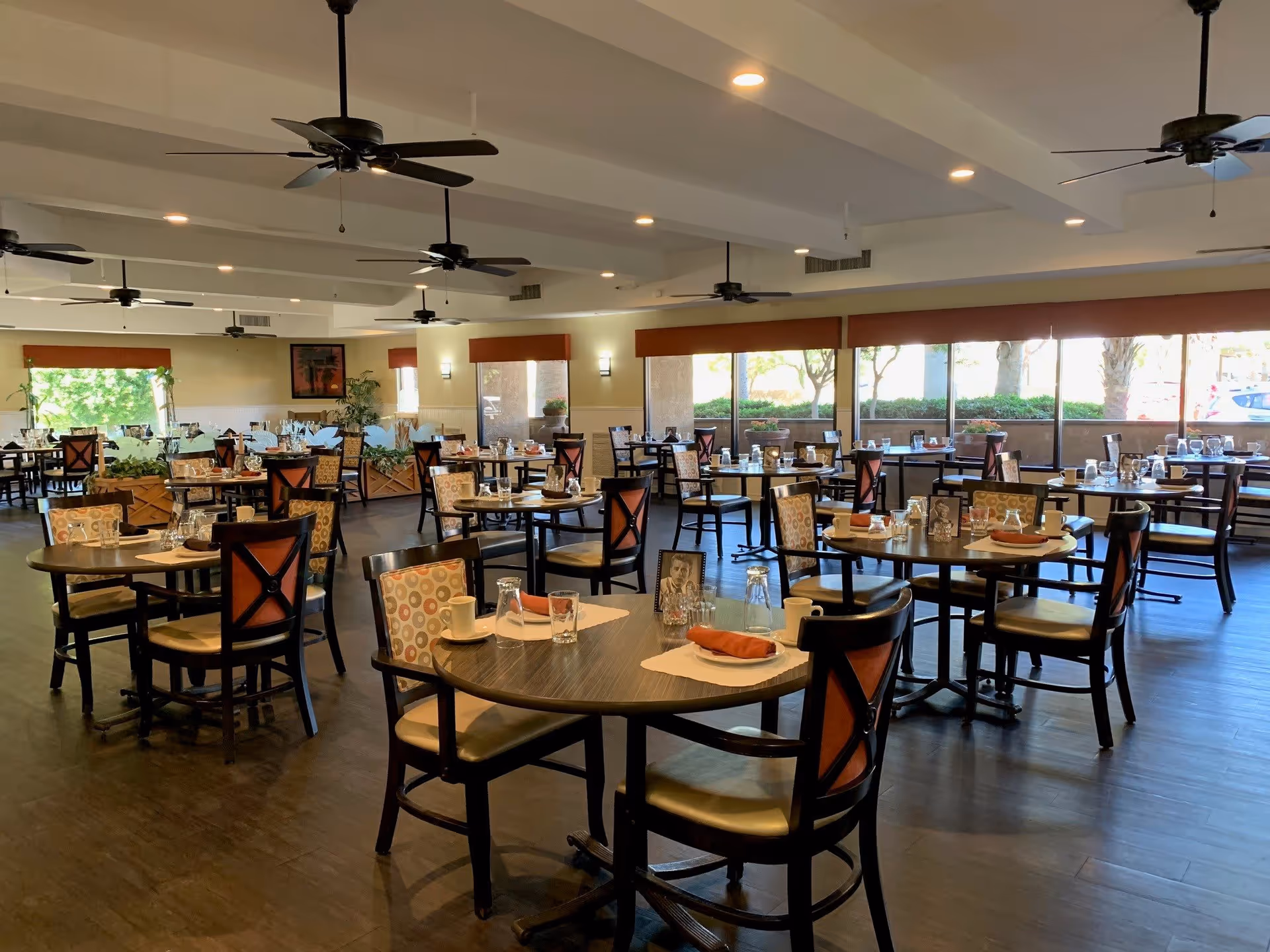 A spacious dining room with multiple round tables set with placemats, glasses, cups, and napkins. The chairs have patterned backs and cushioned seats. Large windows along one wall let in natural light, and ceiling fans hang from the white ceiling. There are plants and framed artwork on the walls, creating a welcoming atmosphere.