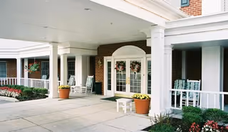 Covered entrance area of a senior living facility with white columns, rocking chairs, potted plants, and flower beds near the doorway.