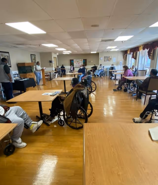 A large room with wooden floors and ceiling lights, filled with elderly individuals seated in wheelchairs around tables. A person stands at a podium speaking to the group. The room has windows with curtains on the right side and some framed pictures on the walls.