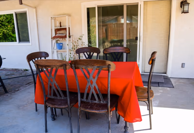 Outdoor patio area with a rectangular table covered by a red tablecloth surrounded by six wooden chairs. The patio is adjacent to a house with sliding glass doors and a window. There is a white shelf with potted plants and other items against the wall.