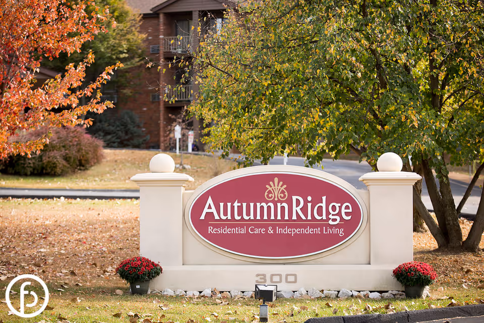 Outdoor view of a sign for Autumn Ridge Residential Care & Independent Living, surrounded by trees with autumn foliage and two potted red flowers on either side of the sign.
