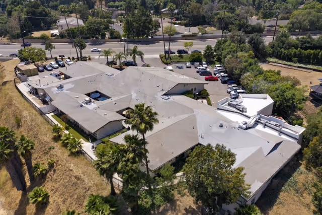 Aerial view of a large single-story building with a gray roof surrounded by trees and vegetation. There is a parking lot with several cars visible near the building, and a road with moving vehicles in the background. The area appears to be suburban with greenery and open spaces around.