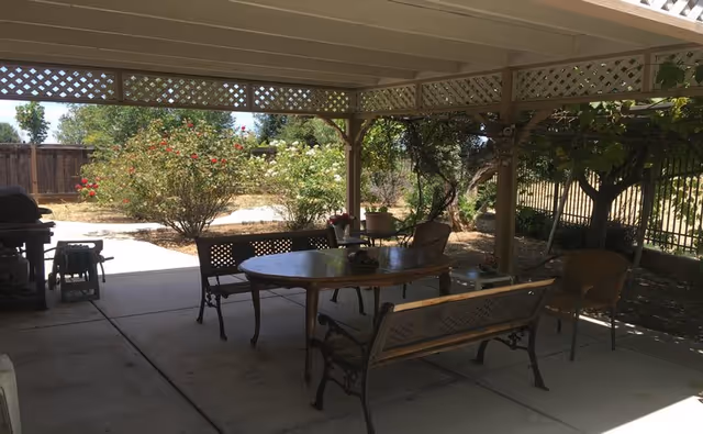 Covered patio with an oval dining table, benches and chairs under a pergola, a grill to the side, and a garden beyond.