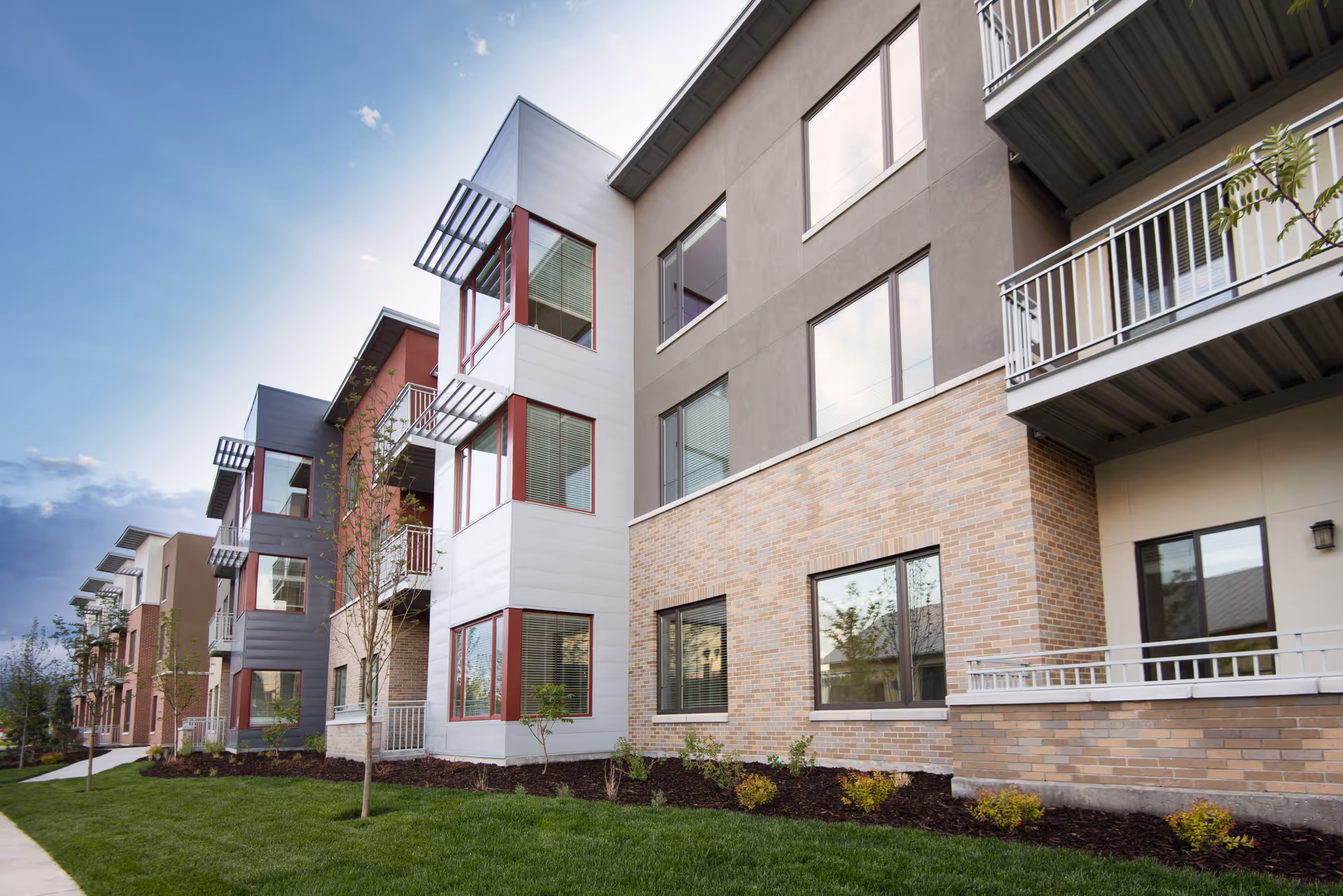 Exterior view of a modern multi-story residential building with large windows, balconies, and a well-maintained lawn with small trees and shrubs under a clear sky.