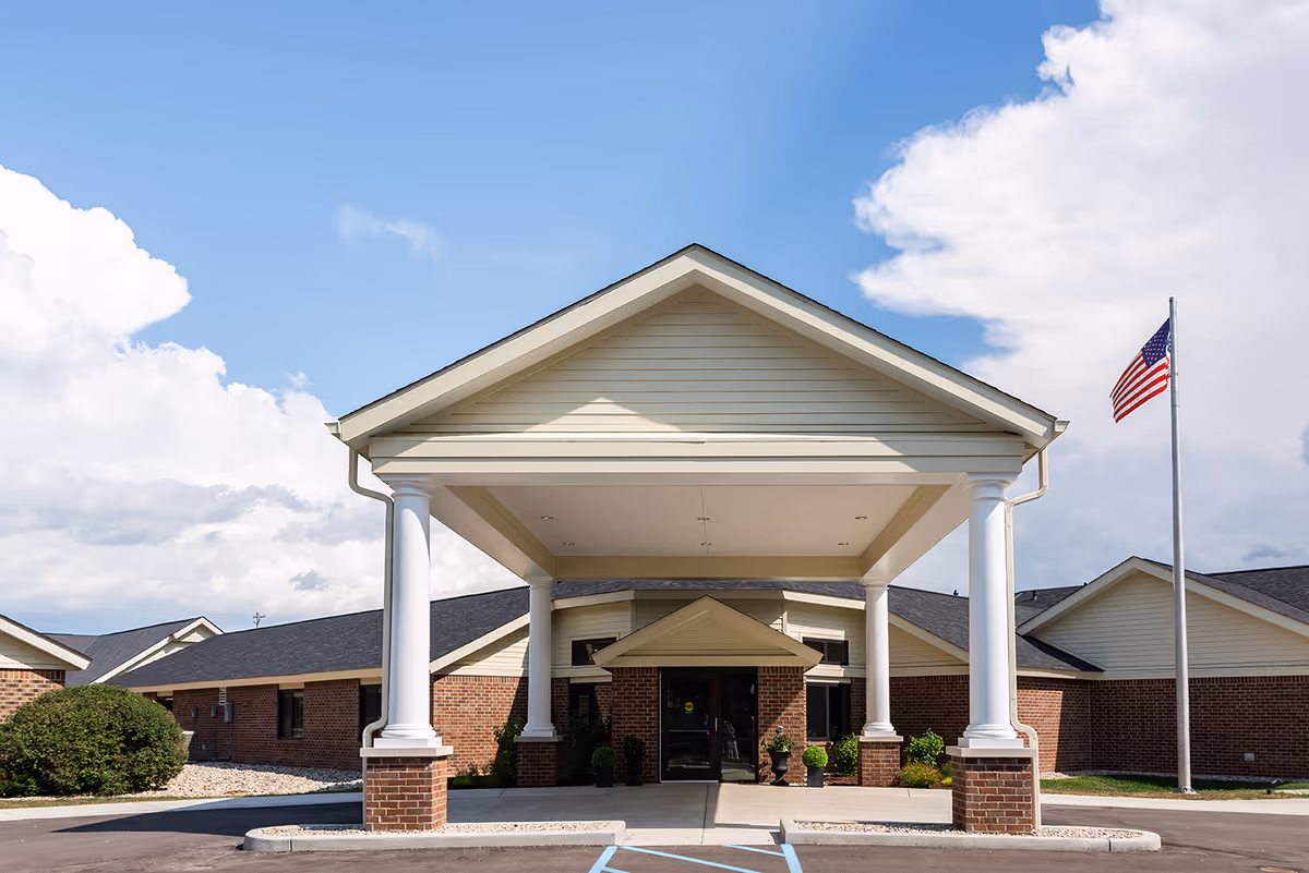 Front exterior view of Rawlins House Family-first Senior Living facility showing a covered entrance with white columns, brick walls, an American flag on a flagpole, and a partly cloudy blue sky.