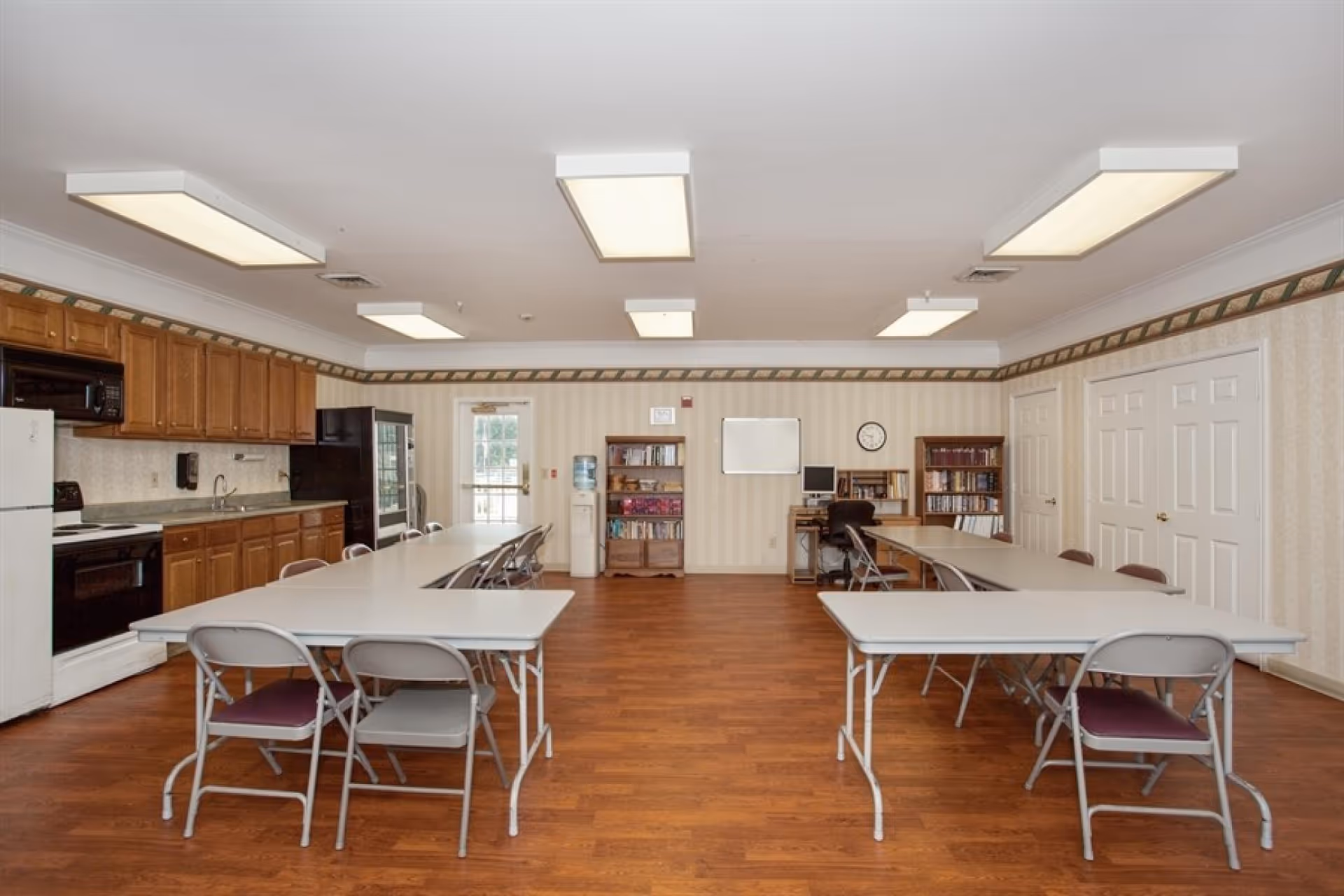 Large communal activity/dining room with folding tables and chairs, a kitchenette on the left, and bookshelves and a computer station along the back wall.