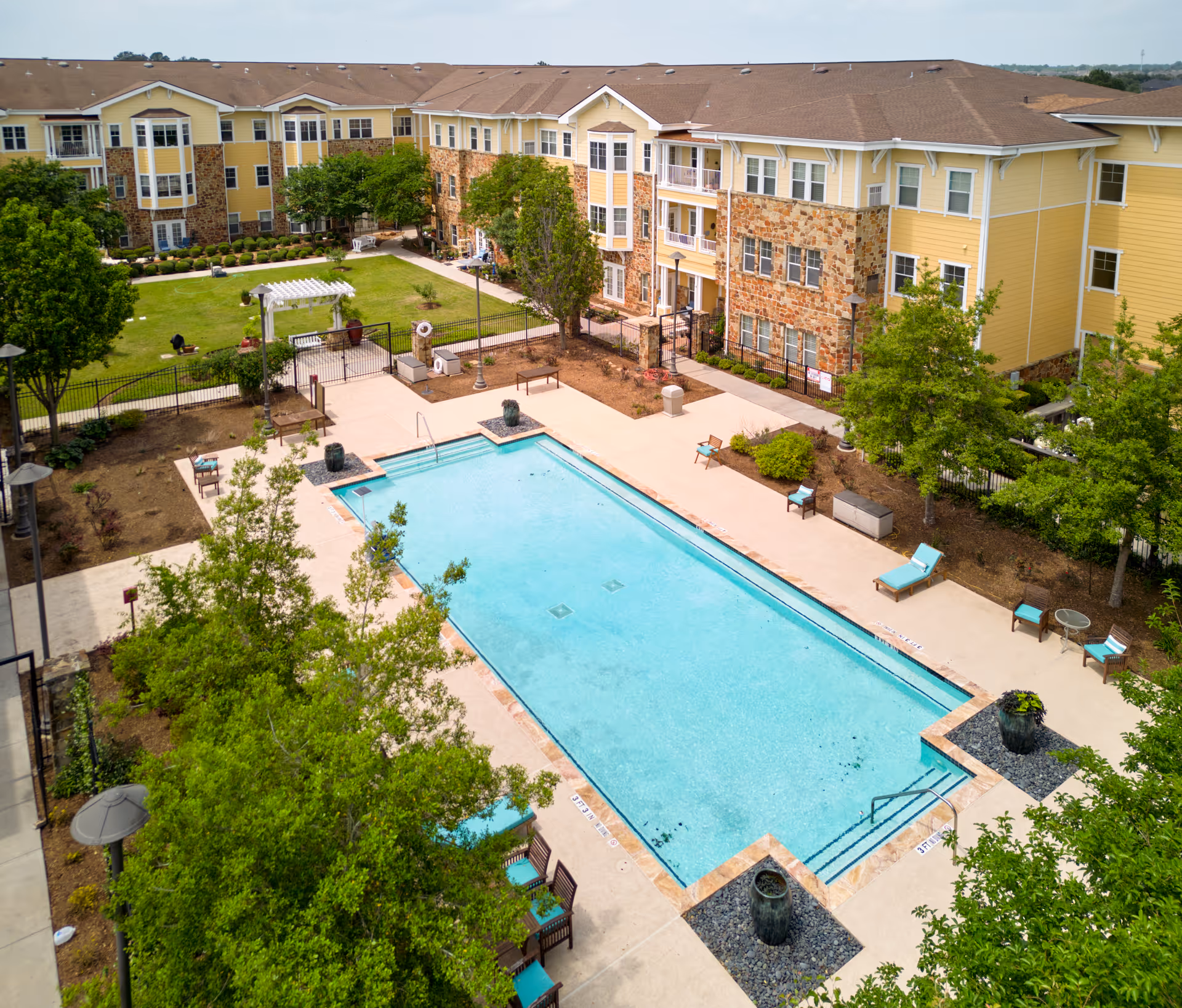 Aerial view of an outdoor swimming pool surrounded by lounge chairs and trees, with a multi-story residential building in the background featuring yellow siding and stone accents.