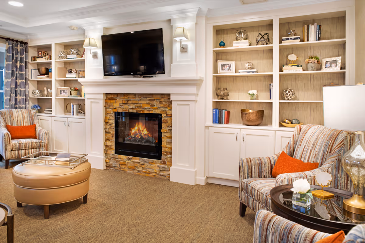 Cozy living room with a stone fireplace and TV between built-in shelves, upholstered armchairs with orange pillows, and a round ottoman.