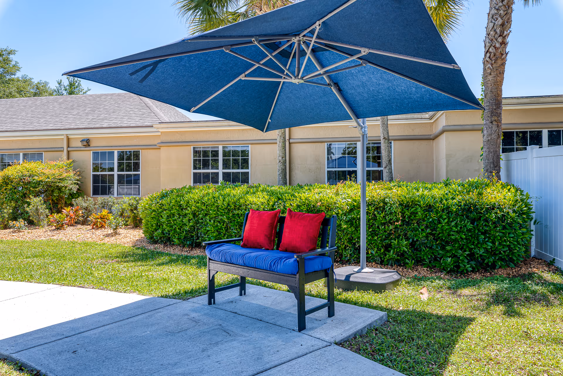 Outdoor bench with a blue cushion and red pillows under a large blue umbrella in front of a single-story building and hedges.