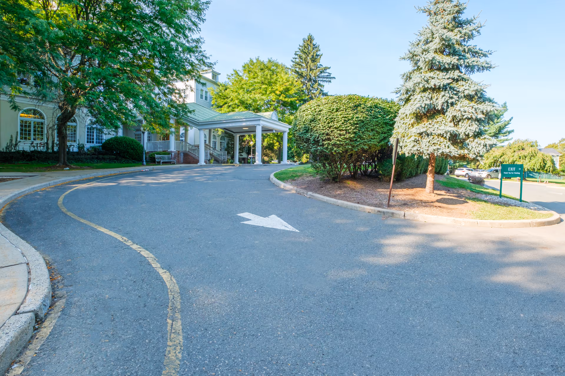 Driveway leading to the entrance of Green Hill Senior Living and Rehabilitation, surrounded by green trees and bushes, with a covered drop-off area and a sign indicating the exit to the right.