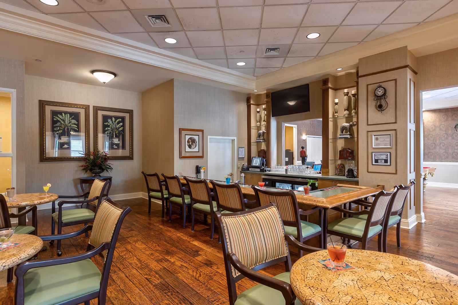 A cozy lounge area with multiple tables and chairs featuring green cushions and striped upholstery. The room has wooden flooring, framed botanical artwork on the walls, and a bar counter with glassware and decorative items. A flat-screen TV is mounted above the bar, and a clock and certificates are displayed on a wooden column. The space is well-lit with ceiling lights and has a warm, inviting atmosphere.
