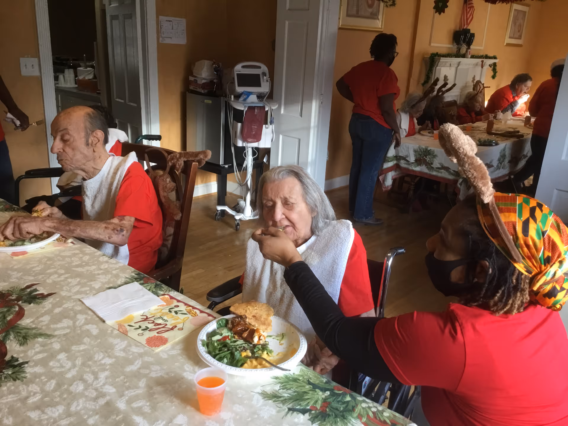 An elderly woman in a wheelchair is being fed by a caregiver wearing a red shirt and a colorful headscarf with reindeer antlers. Another elderly man is seated next to them eating independently. In the background, more elderly residents and caregivers are seated and standing around a dining table decorated with a holiday-themed tablecloth.